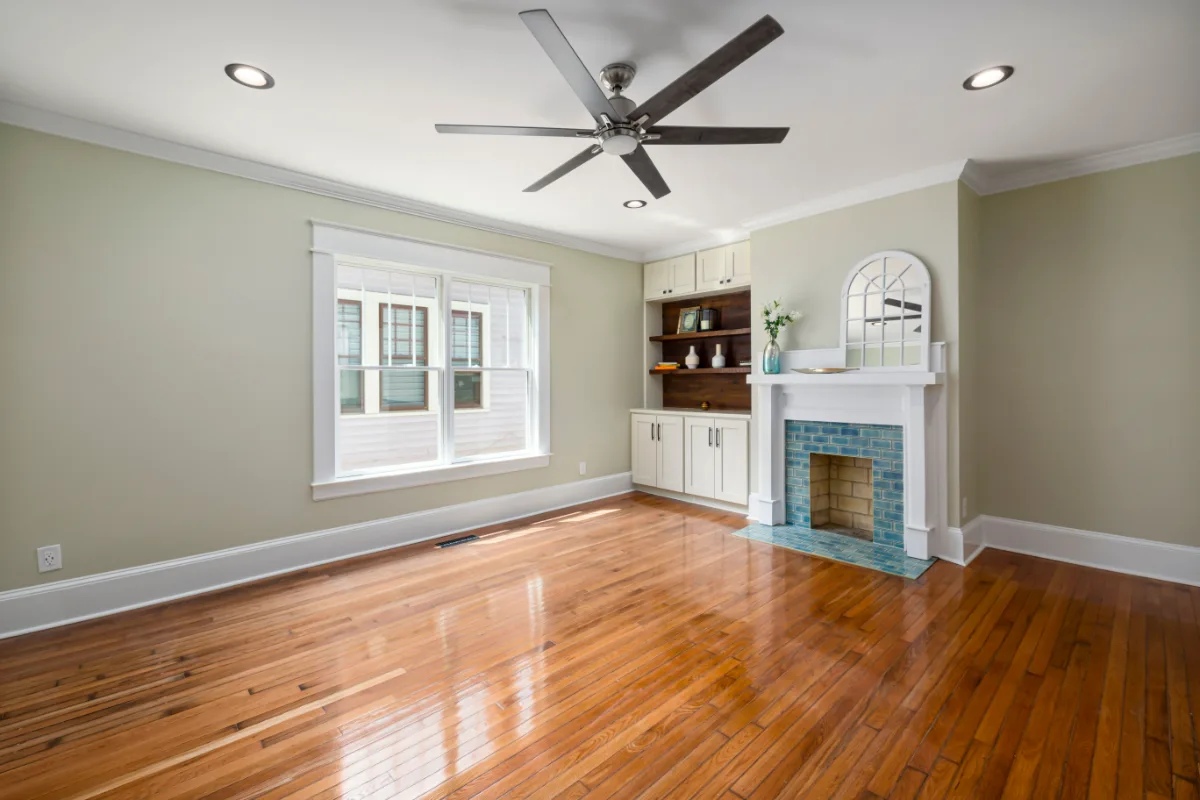 Empty living room with hardwood floors, a tiled fireplace, built-in shelving and a ceiling fan, ready to be virtually staged